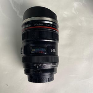 Peut inclure: Une tasse en céramique blanche avec une anse noire et le texte "But First, Coffee" dans un style manuscrit. La tasse est posée sur une surface blanche. La tasse mesure environ 10 cm de haut.