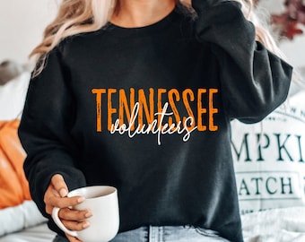 May include: A black sweatshirt with the text "Tennessee Volunteers" in orange and white lettering. The sweatshirt is being worn by a person with long blonde hair. The person is holding a white mug.