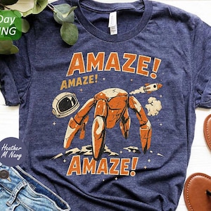 May include: A heather navy blue t-shirt featuring a graphic print of an orange robot on a lunar surface with an astronaut helmet and a small rocket. The text "AMAZE!" appears above and below the robot in a bold, distressed orange font.