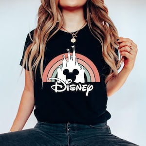 K&ouml;nnte beinhalten: Schwarzes T-Shirt mit einer wei&szlig;en Silhouette von Schloss Aschenputtel und Mickey-Maus-Ohren vor einem Regenbogen. Der Text "DISNEY" steht unterhalb des Schlosses.