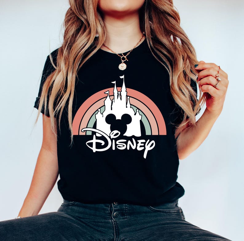 K&ouml;nnte beinhalten: Schwarzes T-Shirt mit einer wei&szlig;en Silhouette von Schloss Aschenputtel und Mickey-Maus-Ohren vor einem Regenbogen. Der Text "DISNEY" steht unterhalb des Schlosses.