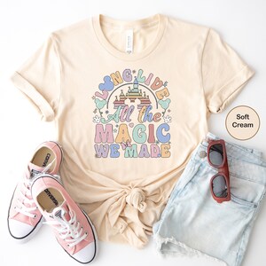 May include: A soft cream colored t-shirt with a rainbow graphic that says "Long Live All the Magic We Made". The graphic features a cartoon castle and Mickey Mouse ears. The shirt is paired with denim shorts and pink Converse sneakers.