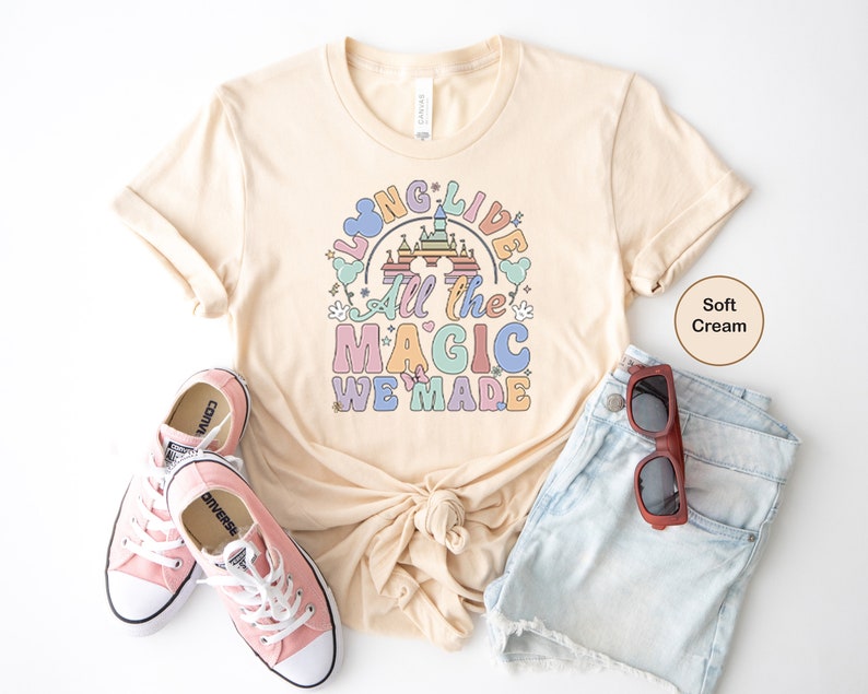 May include: A soft cream colored t-shirt with a rainbow graphic that says "Long Live All the Magic We Made". The graphic features a cartoon castle and Mickey Mouse ears. The shirt is paired with denim shorts and pink Converse sneakers.