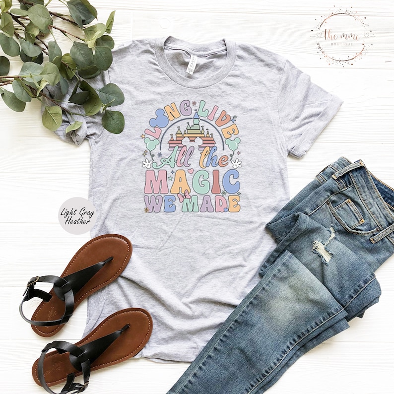 May include: A light gray heather t-shirt with a colorful graphic design featuring a castle, Mickey Mouse ears, and the text "Long Live All the Magic We Made".