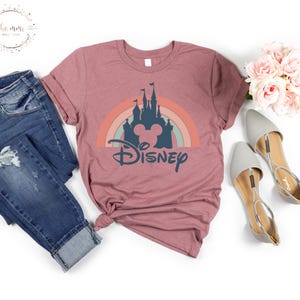 K&ouml;nnte beinhalten: Ein mauvefarbenes T-Shirt mit einem Aufdruck eines Regenbogens, eines Schlosses und Mickey Mauses Kopf. Der Text "DISNEY" befindet sich unter dem Aufdruck.