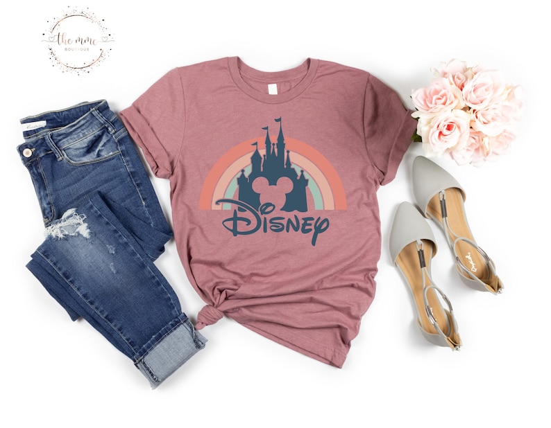 K&ouml;nnte beinhalten: Ein mauvefarbenes T-Shirt mit einem Aufdruck eines Regenbogens, eines Schlosses und Mickey Mauses Kopf. Der Text "DISNEY" befindet sich unter dem Aufdruck.