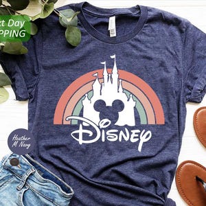 K&ouml;nnte beinhalten: Ein marineblaues T-Shirt mit einem wei&szlig;en Aufdruck eines Regenbogens, eines Schlosses und Mickey Mouse Ohren. Der Text "DISNEY" befindet sich unter dem Aufdruck.