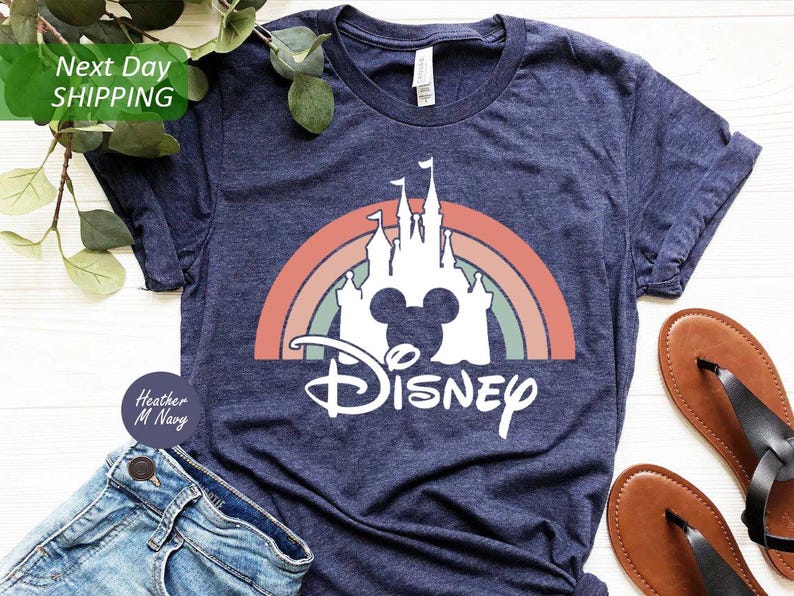 K&ouml;nnte beinhalten: Ein marineblaues T-Shirt mit einem wei&szlig;en Aufdruck eines Regenbogens, eines Schlosses und Mickey Mouse Ohren. Der Text "DISNEY" befindet sich unter dem Aufdruck.
