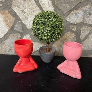 May include: Two ceramic planters shaped like people sitting in a meditative pose. The planter on the left is red and the planter on the right is pink. A small potted plant with green leaves is in the background.
