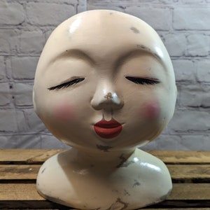 May include: White ceramic bust of a woman with closed eyes and red lips. The bust is painted with pink cheeks and black eyelashes. The bust is sitting on a wooden surface with the word "Mexico" written on it.