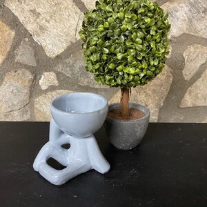 May include: A light blue ceramic planter shaped like a person sitting with legs crossed. The planter has a round bowl on top. A small artificial topiary tree is in a gray pot next to the planter.