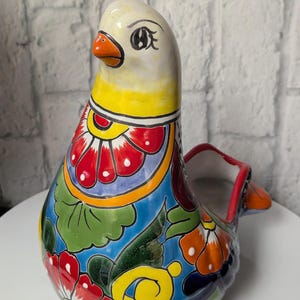 May include: A colorful ceramic bird figurine with a blue, green, yellow, red, and white floral pattern. The bird has a white head with a yellow beak and black eyes.