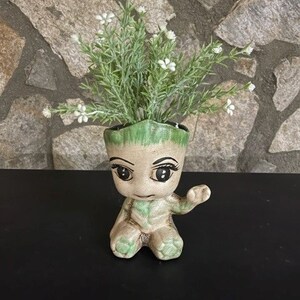 May include: A ceramic planter shaped like a green and brown character with large eyes and a friendly expression. The planter is holding a small green plant with white flowers.