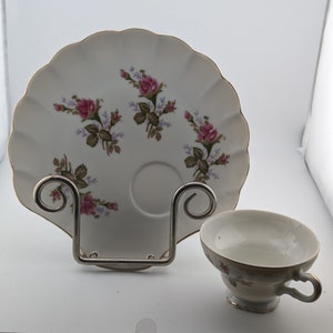 May include: A white teacup and saucer set with a delicate floral pattern featuring pink roses and green leaves. The saucer has a scalloped edge and a gold rim.