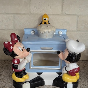 May include: A blue and white ceramic cookie jar shaped like a vintage oven with Mickey Mouse and Minnie Mouse figurines on either side.  Goofy is on top of the oven, peeking into the oven window.  The oven has a black and white checkered base.
