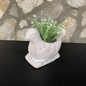May include: A white ceramic bird planter with a small plant with white flowers inside. The planter is sitting on a black surface.