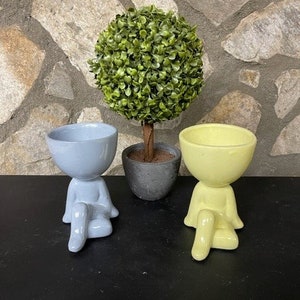 May include: Two ceramic planters shaped like people sitting with legs crossed. The planter on the left is light blue and the planter on the right is yellow. A small green topiary tree is in the background.