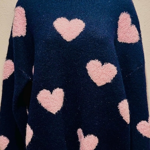 Puede incluir: Suéter azul marino con grandes corazones rosa claro. El suéter tiene cuello redondo y mangas largas. Los corazones son de un material suave y texturizado, que contrasta con el punto liso del suéter. Un estilo casual y cómodo.