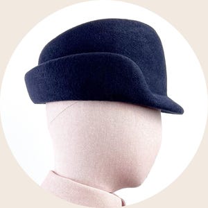 May include: A navy blue felt hat with a rounded crown and a brim that curves around the forehead. The hat is displayed on a mannequin head against a white background.