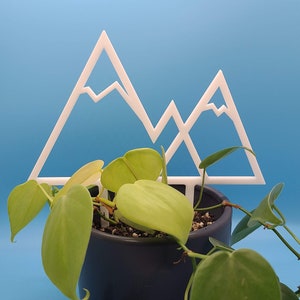 May include: A white plastic mountain-shaped plant stake is in a dark gray pot with a green leafy plant growing around it.