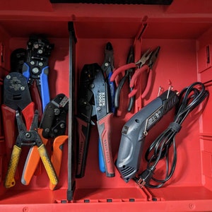 May include: A red toolbox filled with various electrical tools. The tools include wire strippers, crimpers, pliers, and a heat gun. The tools have black, red, blue, and yellow handles. The toolbox is open, revealing the organized arrangement of the tools.