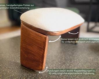 May include: A wooden footrest with a white padded top. The legs fold for easy storage and transport. The footrest is designed to tilt slightly for ergonomic seating.