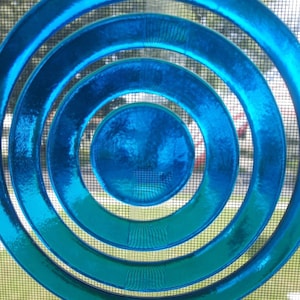 May include: A circular suncatcher featuring a series of concentric rings in varying shades of blue, ranging from a deep cyan to a lighter sky blue. The glass has a textured, rippled surface that creates light refraction and depth across the design.