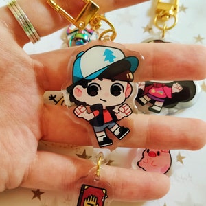 May include: A gold keychain with a clear charm featuring a cartoon character with a blue hat and red shirt. The charm is attached to a smaller charm with a red book with a hand and the number 3 on the cover.