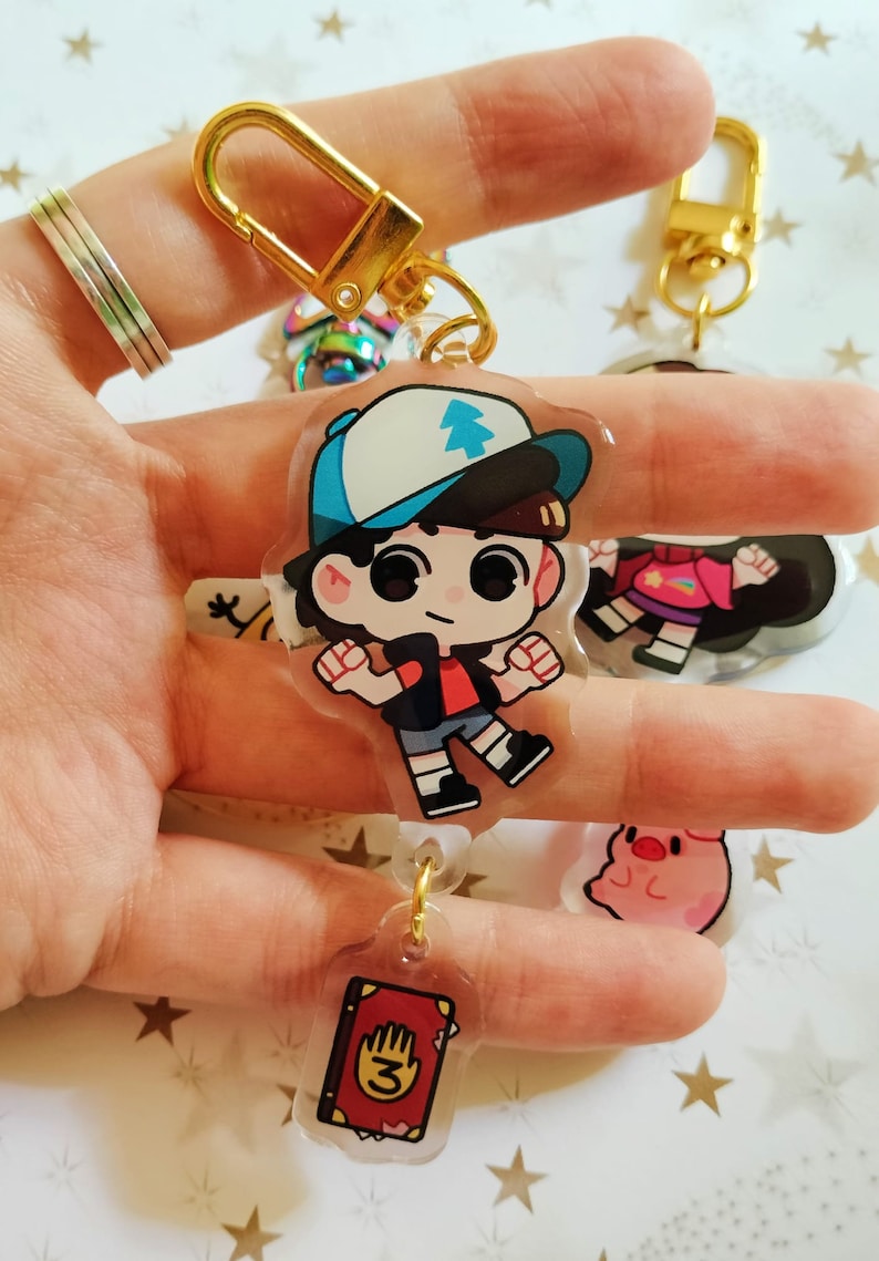 May include: A gold keychain with a clear charm featuring a cartoon character with a blue hat and red shirt. The charm is attached to a smaller charm with a red book with a hand and the number 3 on the cover.