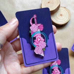 May include: A pink and green enamel pin featuring a sword with pink roses and a pink gem. The pin is on a purple card with white clouds and the text "@ketsutsuki".