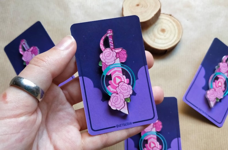 May include: A pink and green enamel pin featuring a sword with pink roses and a pink gem. The pin is on a purple card with white clouds and the text "@ketsutsuki".