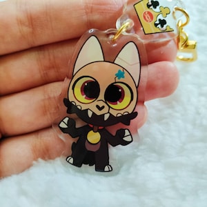 May include: A clear acrylic keychain featuring a cartoon character with a black and white fur pattern, yellow eyes, and a blue star on its forehead. The character is wearing a red collar with a gold tag.