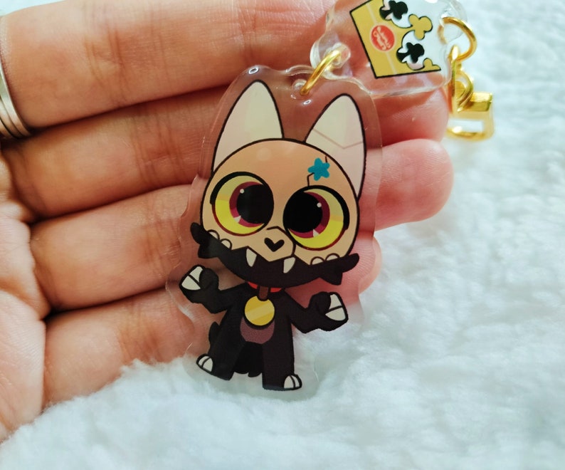 May include: A clear acrylic keychain featuring a cartoon character with a black and white fur pattern, yellow eyes, and a blue star on its forehead. The character is wearing a red collar with a gold tag.