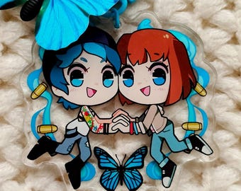 May include: A clear acrylic charm featuring two cartoon characters holding hands, with blue and gold accents. A blue butterfly and a blue keychain are also visible. The charm is on a white, textured surface.