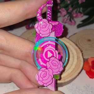 May include: Pink and blue iridescent acrylic charm featuring a sword with roses and a pink gem with a play button inside.
