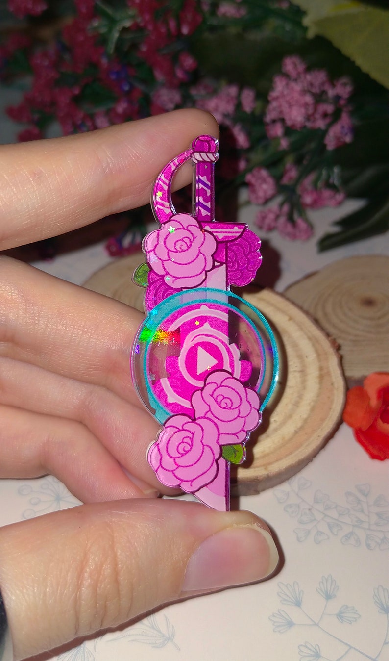 May include: Pink and blue iridescent acrylic charm featuring a sword with roses and a pink gem with a play button inside.