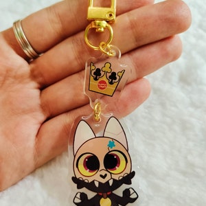 May include: A gold keychain with a clear acrylic charm featuring a cartoon character with a crown. The character has black fur, a gold collar, and a blue star on its forehead. The crown is gold with black card suits.