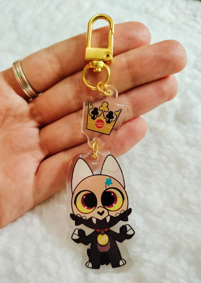 May include: A gold keychain with a clear acrylic charm featuring a cartoon character with a crown. The character has black fur, a gold collar, and a blue star on its forehead. The crown is gold with black card suits.