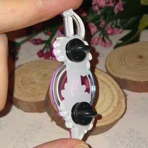 May include: A white and pink enamel pin with a sword design. The pin has a black butterfly on the back and a safety clasp.