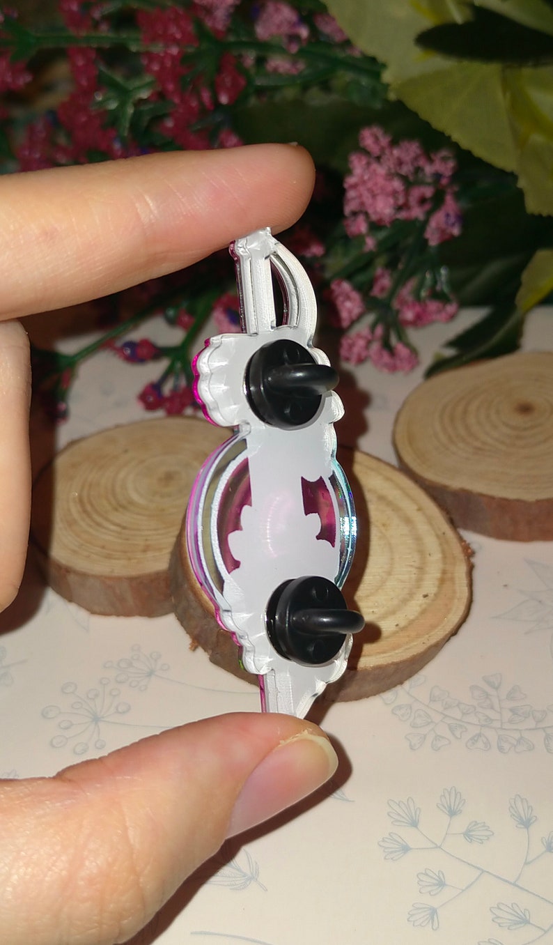May include: A white and pink enamel pin with a sword design. The pin has a black butterfly on the back and a safety clasp.