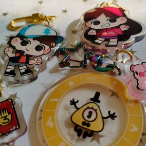 May include: A set of five Gravity Falls themed keychains. The keychains feature characters from the show, including Dipper Pines, Mabel Pines, and Bill Cipher. The keychains are made of clear acrylic and have a colorful design.
