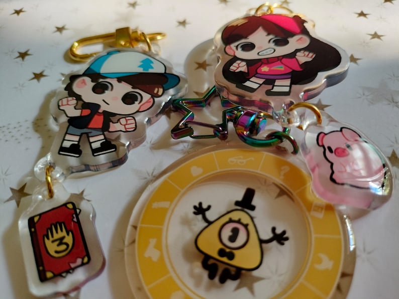 May include: A set of five Gravity Falls themed keychains. The keychains feature characters from the show, including Dipper Pines, Mabel Pines, and Bill Cipher. The keychains are made of clear acrylic and have a colorful design.