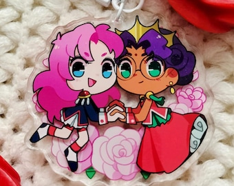 May include: A pink and purple anime character keychain with a white background. The characters are holding hands and are surrounded by pink roses. The keychain has a silver metal clasp.