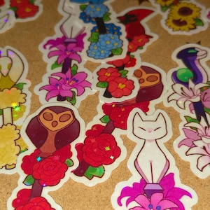 Puede incluir: Una colección de pegatinas coloridas con varios diseños. Las pegatinas incluyen arreglos florales, figuras de gatos, un búho y otras ilustraciones caprichosas. Las pegatinas tienen un borde blanco y están dispuestas sobre una superficie marrón claro.