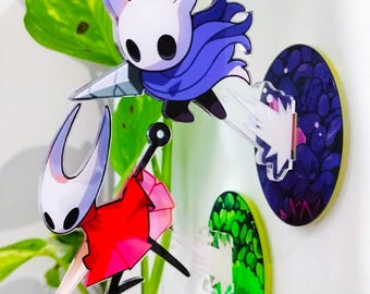 Puede incluir: Acrílico charms con personajes del videojuego Hollow Knight. Uno tiene cabeza blanca y capa azul, el otro cabeza blanca y capa roja. Se adjuntan a bases circulares coloridas. Fondo con follaje verde.