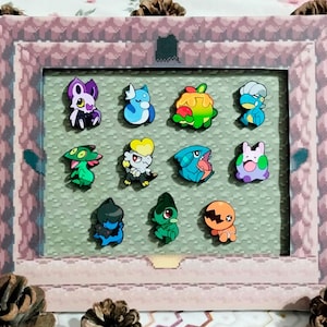 May include: A framed collection of colourful cartoon character pins. The pins are arranged on a green background within a pink and grey pixelated frame. The characters feature various colours and designs, creating a playful and nostalgic aesthetic.