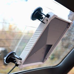 Könnte beinhalten: Ein weißer und grauer Autofensterhalter mit zwei Saugnäpfen. Der Halter ist an einer Heckscheibe eines Autos befestigt.