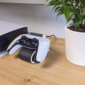 以下が含まれることがあります： 白と黒のPlayStation 5コントローラーが、白と黒の充電スタンドに置かれています。 コントローラーは、白いコンソールと鉢植えの植物の隣の木製の表面にあります。