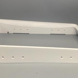 Może przedstawiać: Biała, prostokątna rama z fazowaną krawędzią. Rama ma kilka małych, równomiernie rozmieszczonych otworów wzdłuż boków. Obiekt wykonany jest z litego, nieprzezroczystego materiału i umieszczony na neutralnym szarym tle.