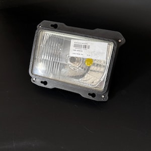May include: A rectangular car headlight with a clear lens and a black plastic frame. A sticker with text is affixed to the lens. The headlight is designed for a Ford Probe 24V.